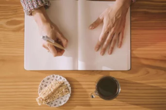 woman's hands with a pen placed on the blank white page next to the cookie and coffee